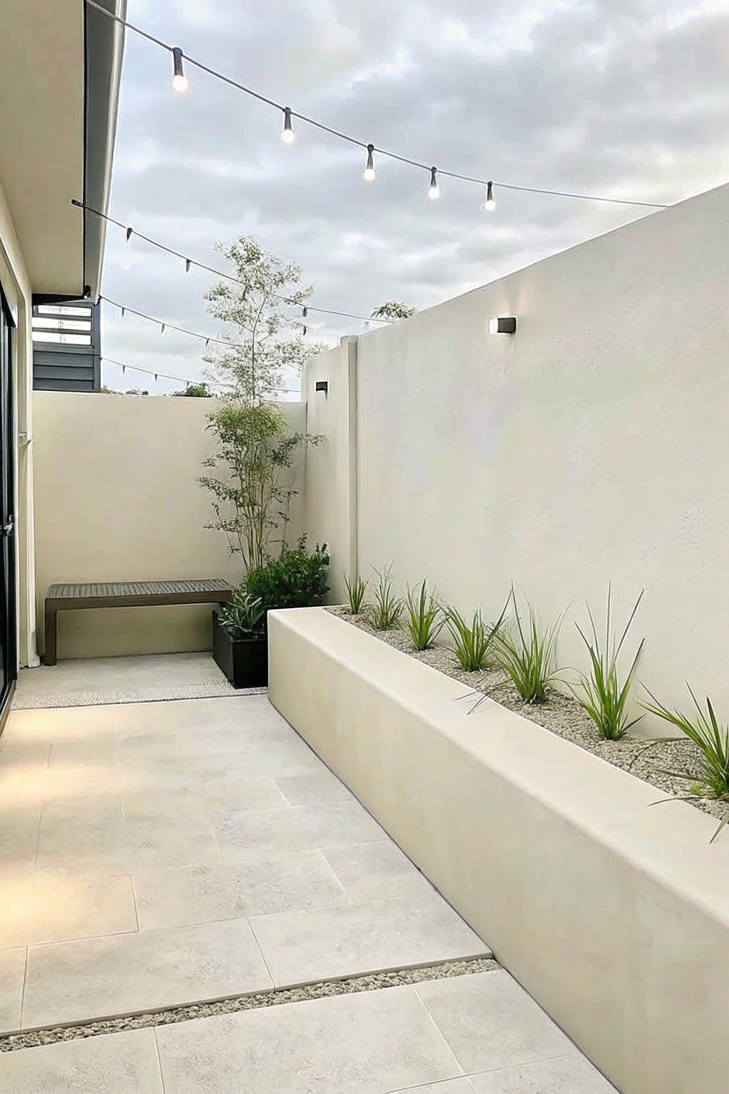 A narrow paved outdoor space with a long raised concrete planter filled with gravel and ornamental grasses running along a white wall.