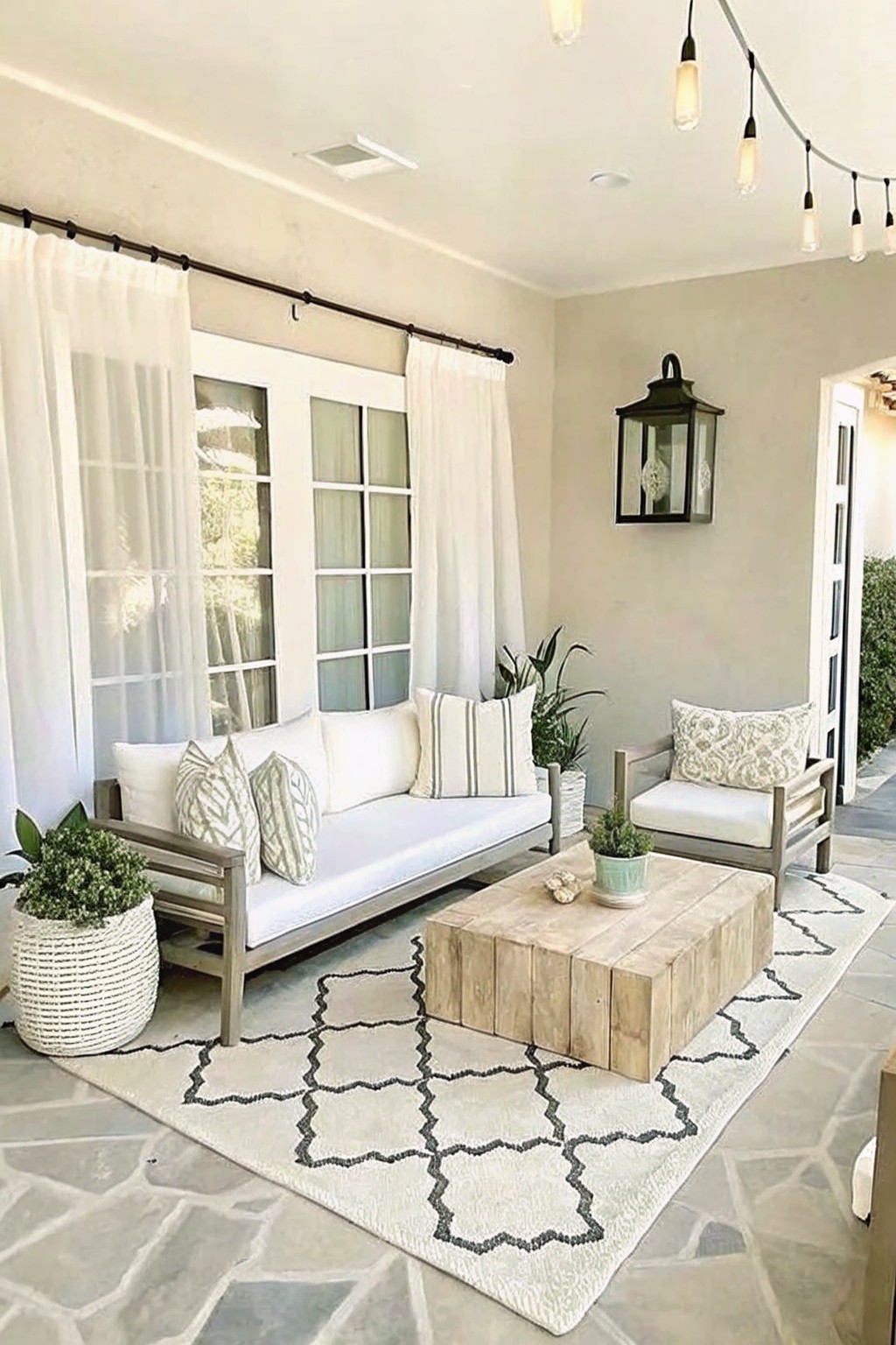 An outdoor patio with a gray sofa, wooden coffee table, patterned rug, and several potted plants in baskets and containers on a stone floor.