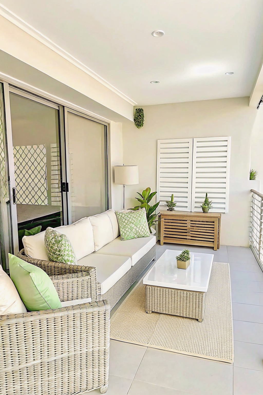 An outdoor patio with white wicker furniture, a white coffee table on a beige rug, a wooden console holding several potted plants, and sliding glass doors on the left.
