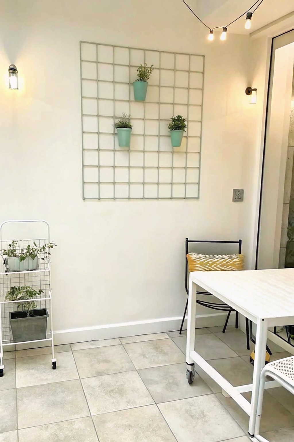 A light-colored patio wall features a large rectangular metal grid holding three small mint green planters, with a white rolling plant cart, table, and chair visible nearby.