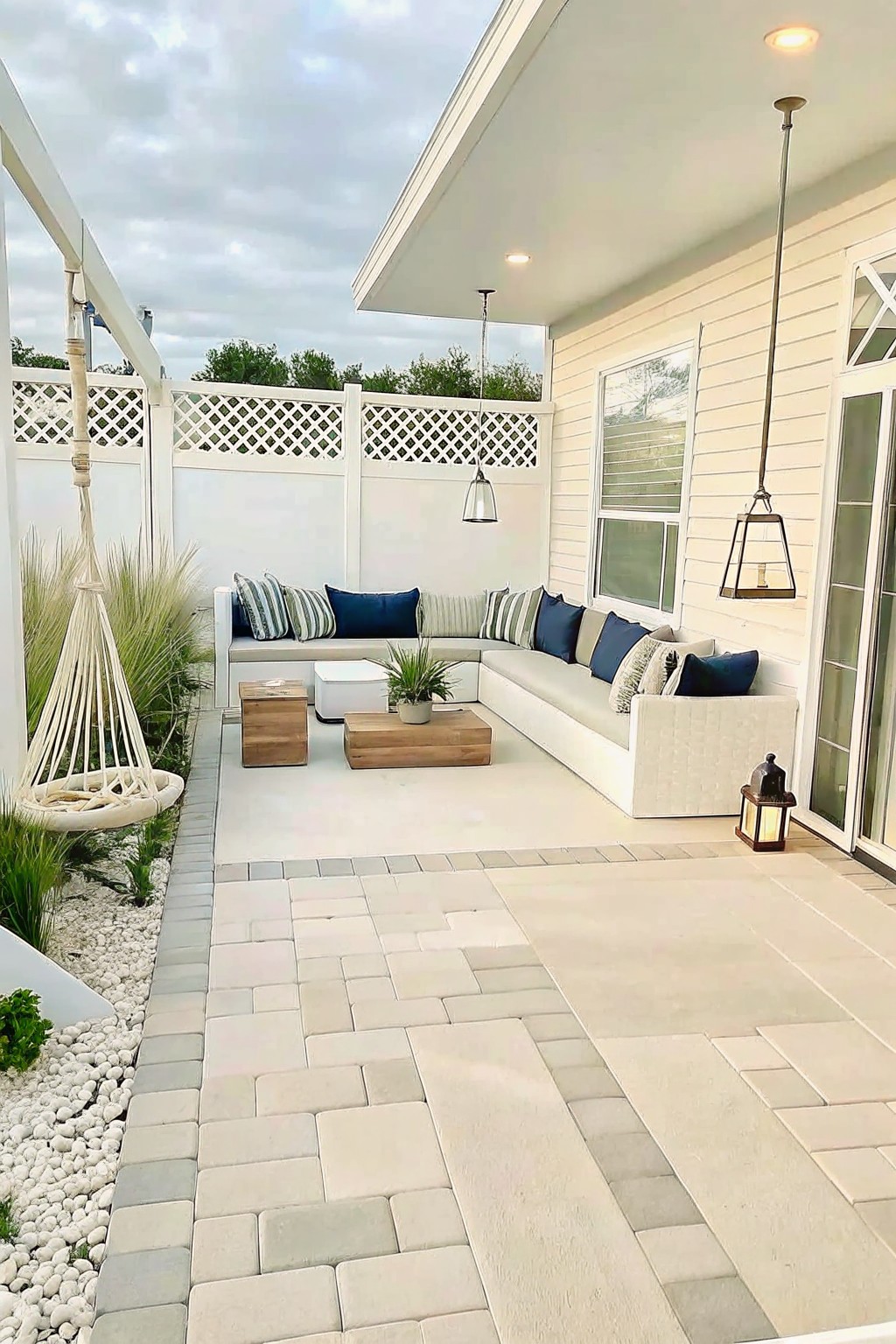 An outdoor patio covered with light gray pavers, featuring an L-shaped sectional sofa with blue and striped cushions, two wooden tables, hanging lights, and a hanging chair beside white fencing.