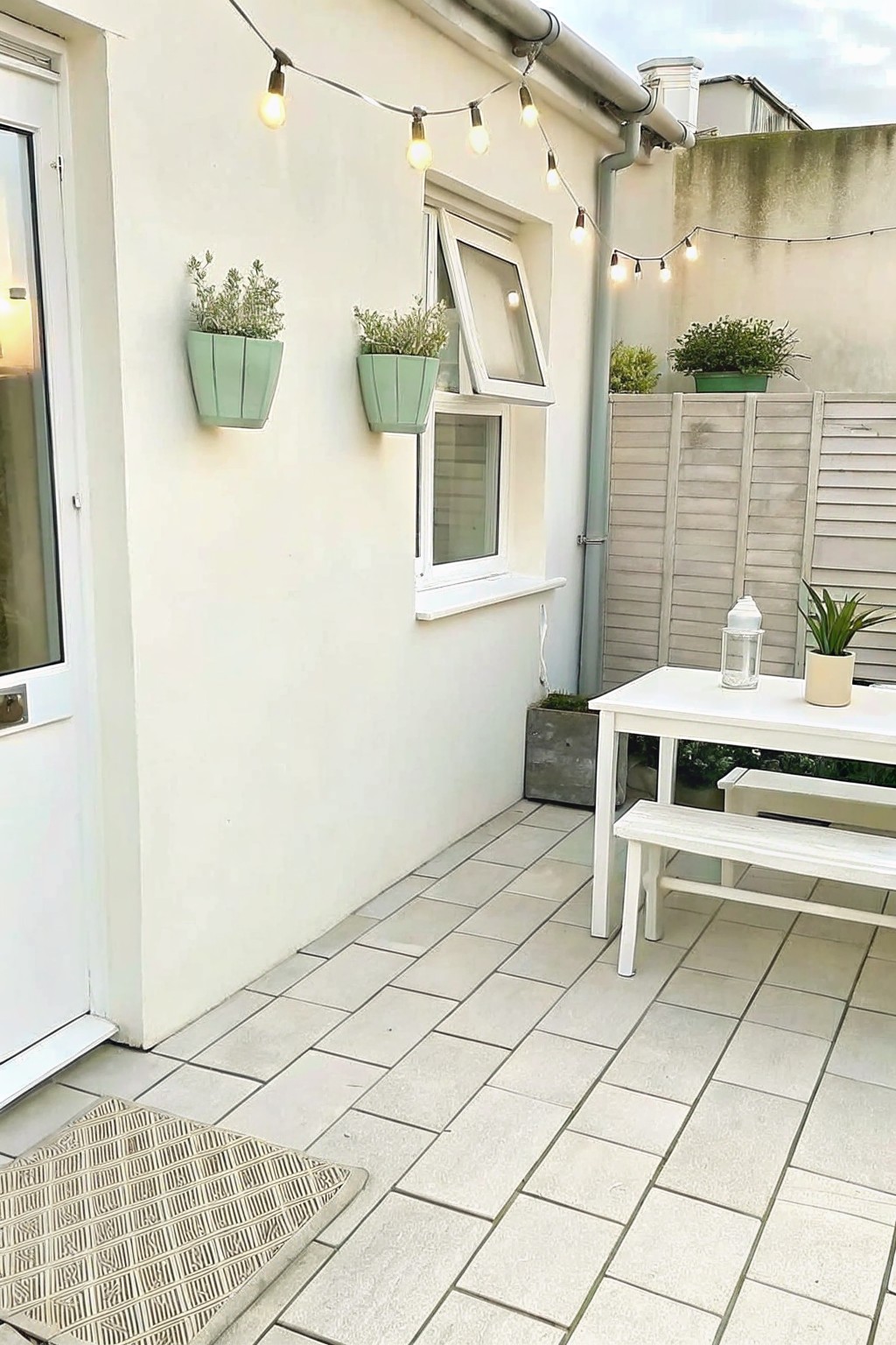 A compact paved patio with a white table and bench set against a house wall that holds two light green mounted planters and string lights overhead.
