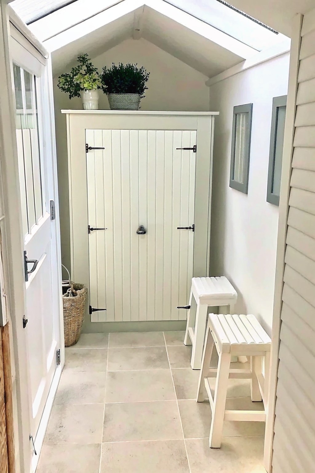 Interior of a small white garden shed with a tall built-in cabinet, two stools, and potted plants on top.