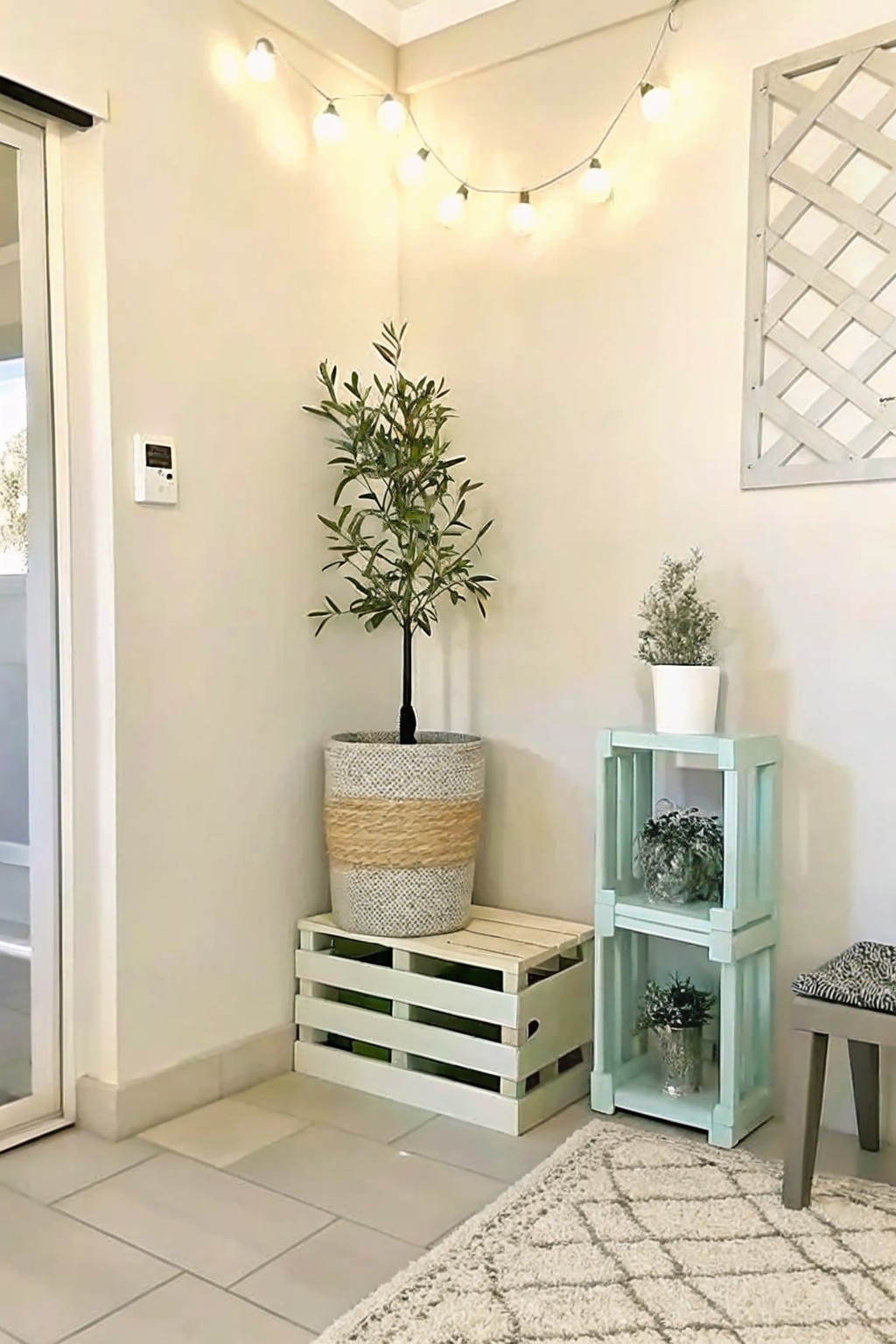 An indoor corner with a large potted tree in a woven basket on a low wooden pallet and two stacked mint-green painted crates holding smaller potted plants.