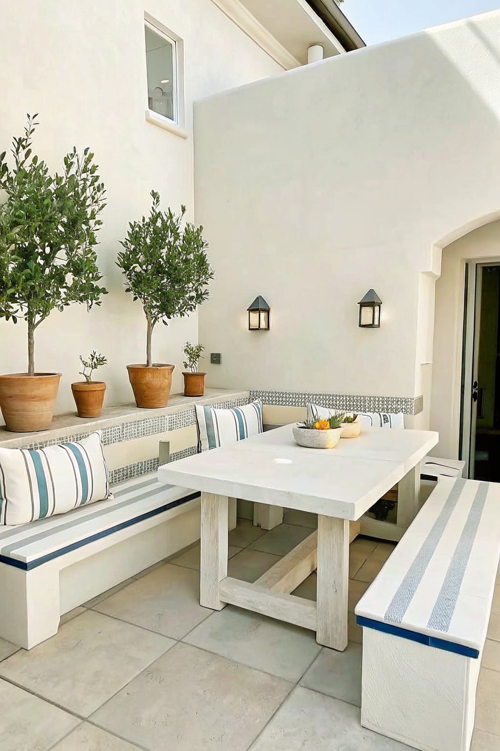 Outdoor patio with white built-in benches featuring blue and gray stripes, a concrete dining table, and terracotta pots with trees and plants along a tiled ledge against a white wall.
