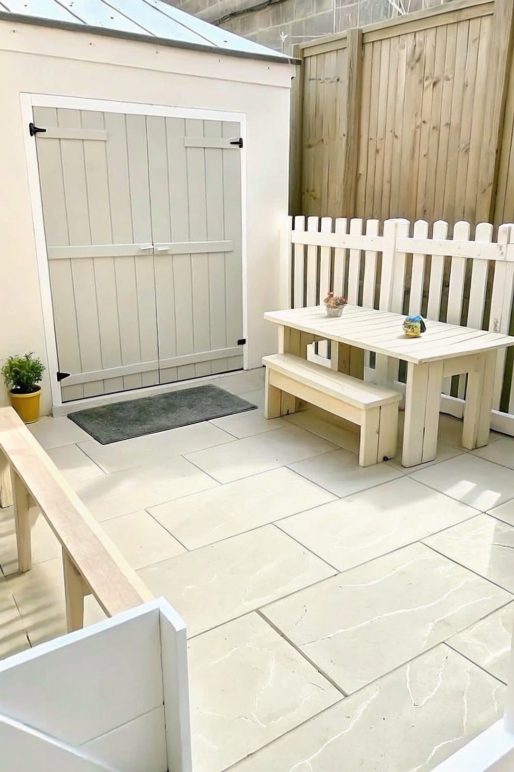 A light gray painted garden shed with double doors beside a white picket fence and wooden picnic table on a tiled patio.