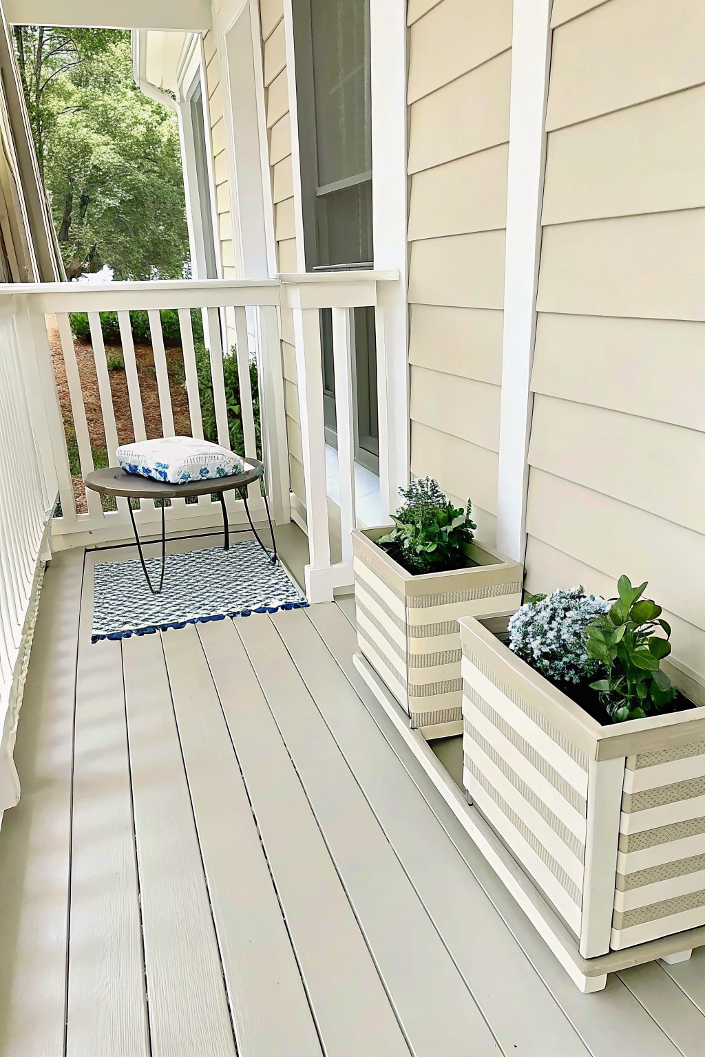 A light gray wooden porch with white railing shows two rectangular planters painted with white and beige stripes holding green plants, next to a small round table, patterned rug, and cushion.