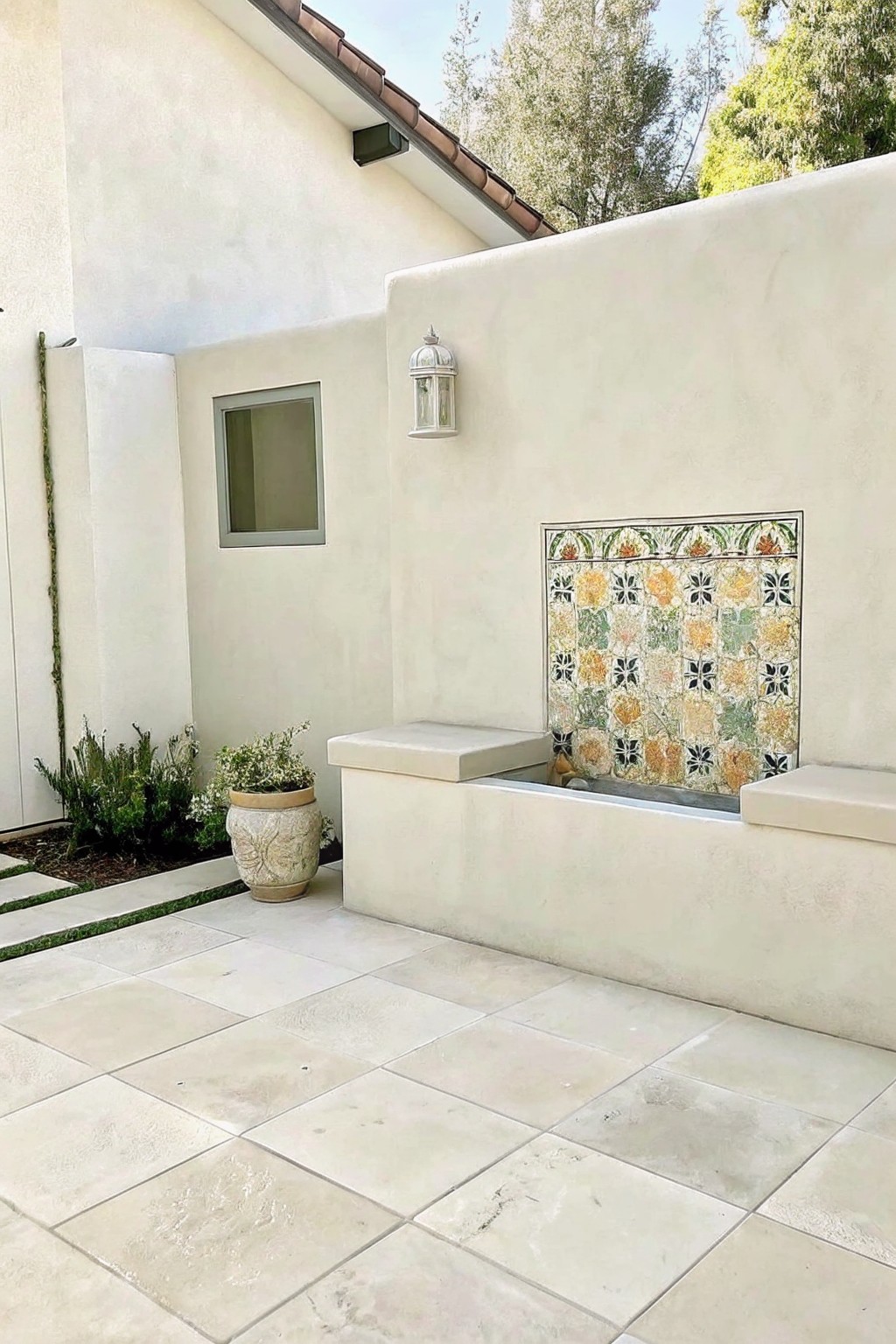 A stucco patio wall with a square colorful tile panel above a small built-in fountain basin, flanked by two concrete benches on a light stone tile floor.