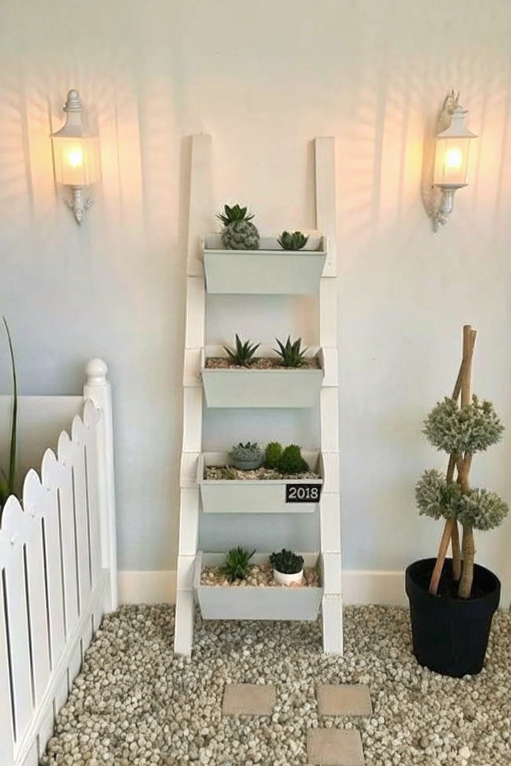 A white wooden ladder repurposed as a multi-tier plant stand holding several small succulent planters, set on gravel outdoors between two wall lanterns.