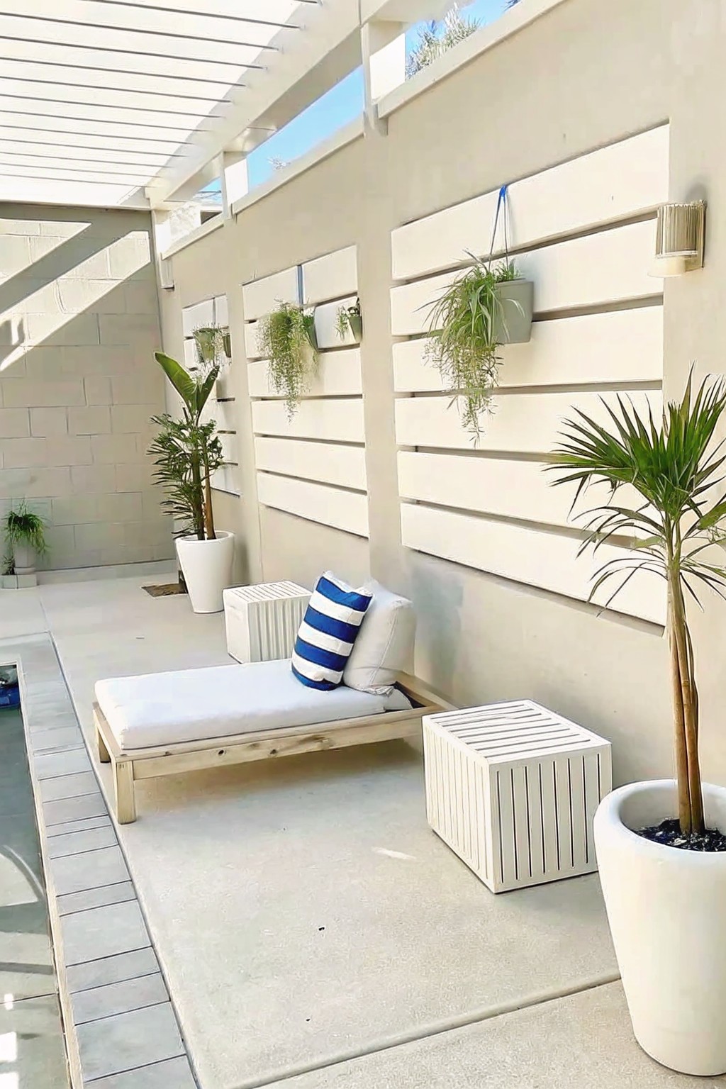 An outdoor patio features a wooden daybed with white cushions and striped pillows, white slatted side tables, large potted plants, and hanging planters on a white horizontal slat wall.