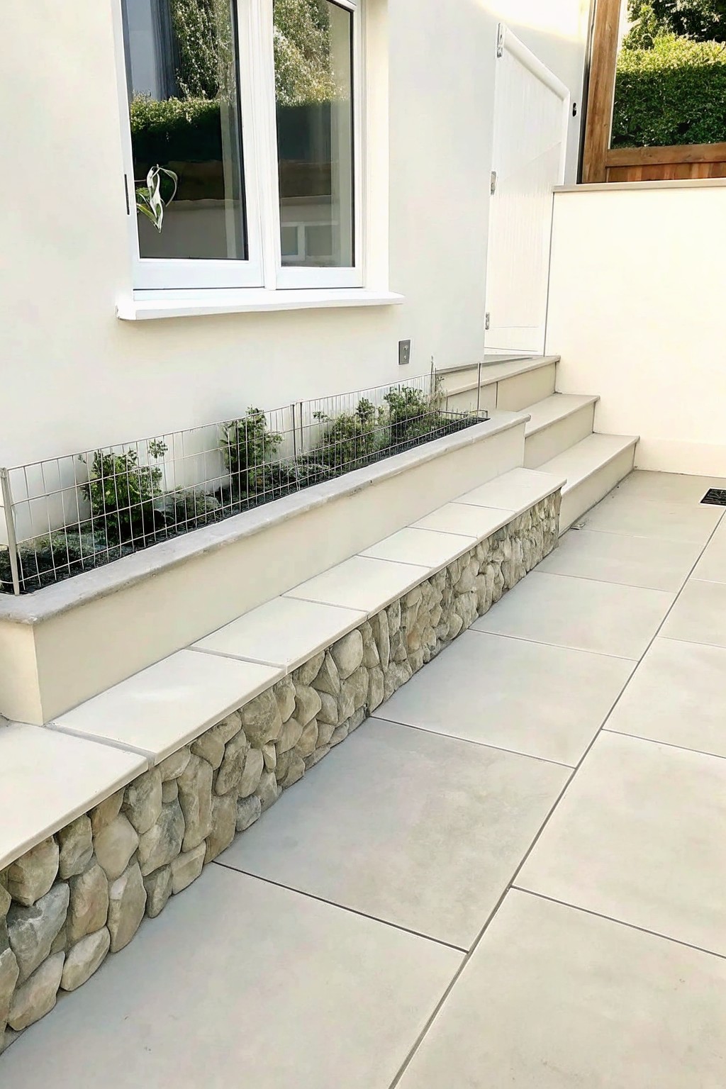 A long raised garden bed with wire mesh and stone sides sits beside concrete steps on a light tiled patio.