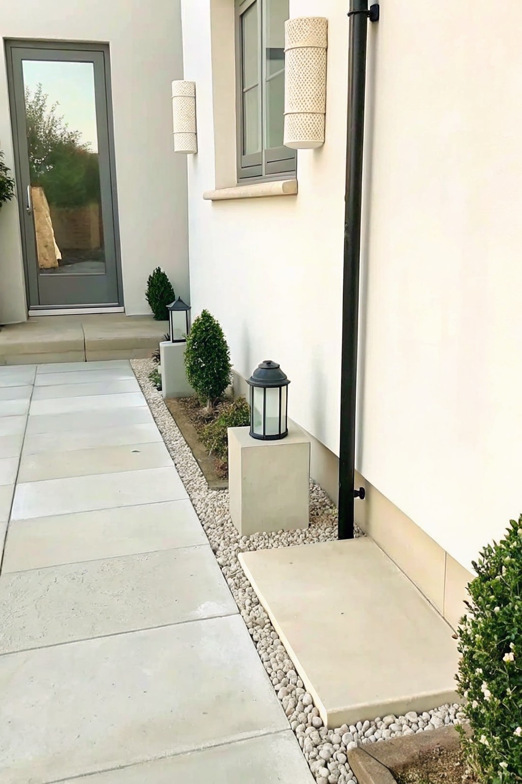 A narrow side yard path of large concrete pavers bordered by white gravel, with small shrubs and two lanterns on concrete blocks next to a white house wall.
