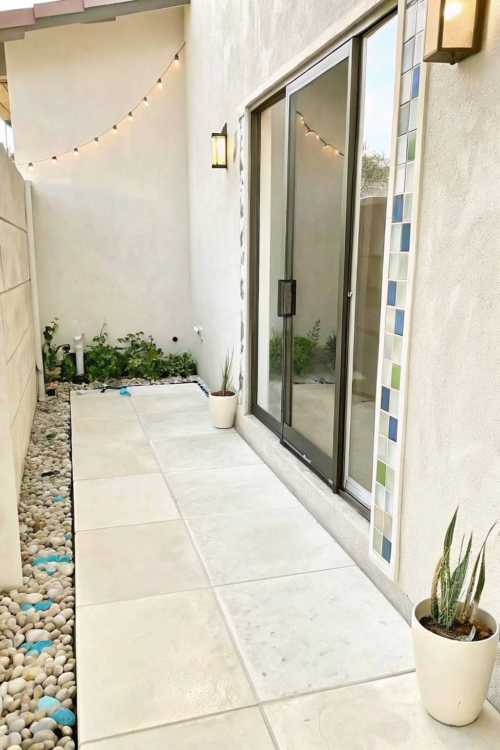 A narrow tiled walkway bordered by a strip of pebbles and low green plants, next to a house with a sliding glass door and a potted succulent in the foreground.