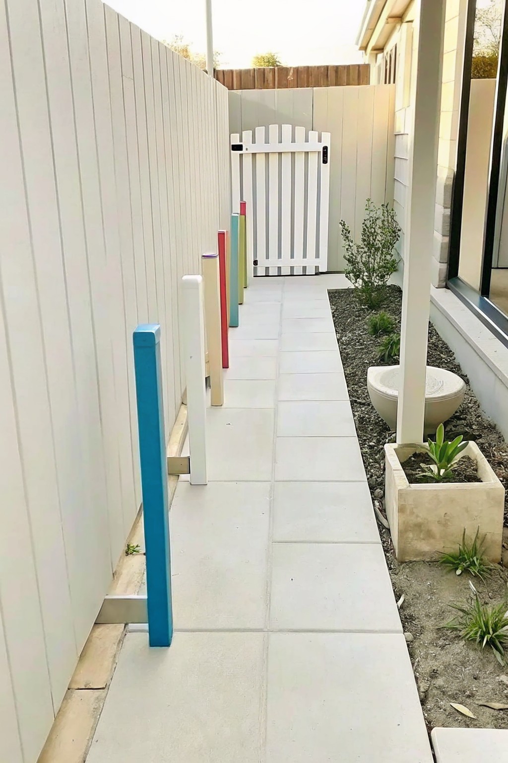 A narrow concrete walkway runs between a white fence and a row of short multicolored painted wooden posts, with a gravel garden bed and concrete planter on the right side.