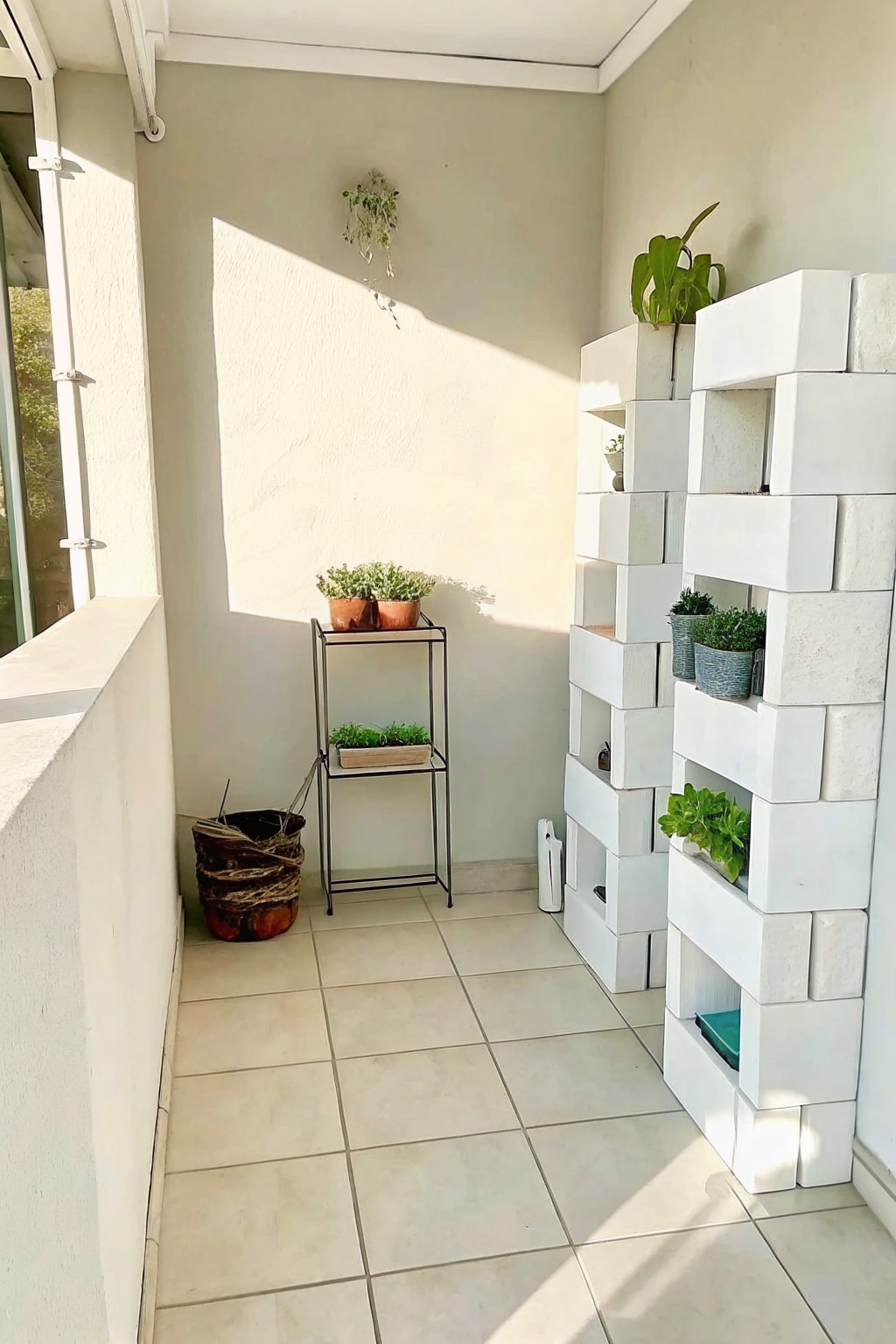Sunlit balcony with white cinder block shelves holding potted plants and metal stand.
