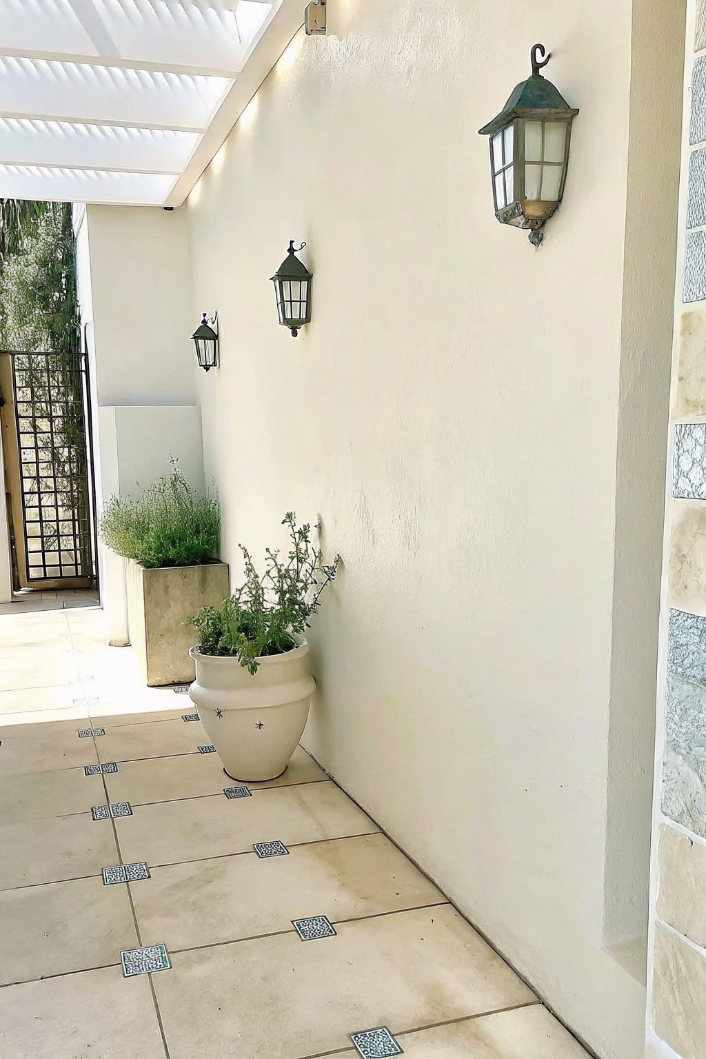 Outdoor covered walkway with a white wall, three dark metal lanterns mounted at staggered heights, and two large potted plants on a tiled floor.