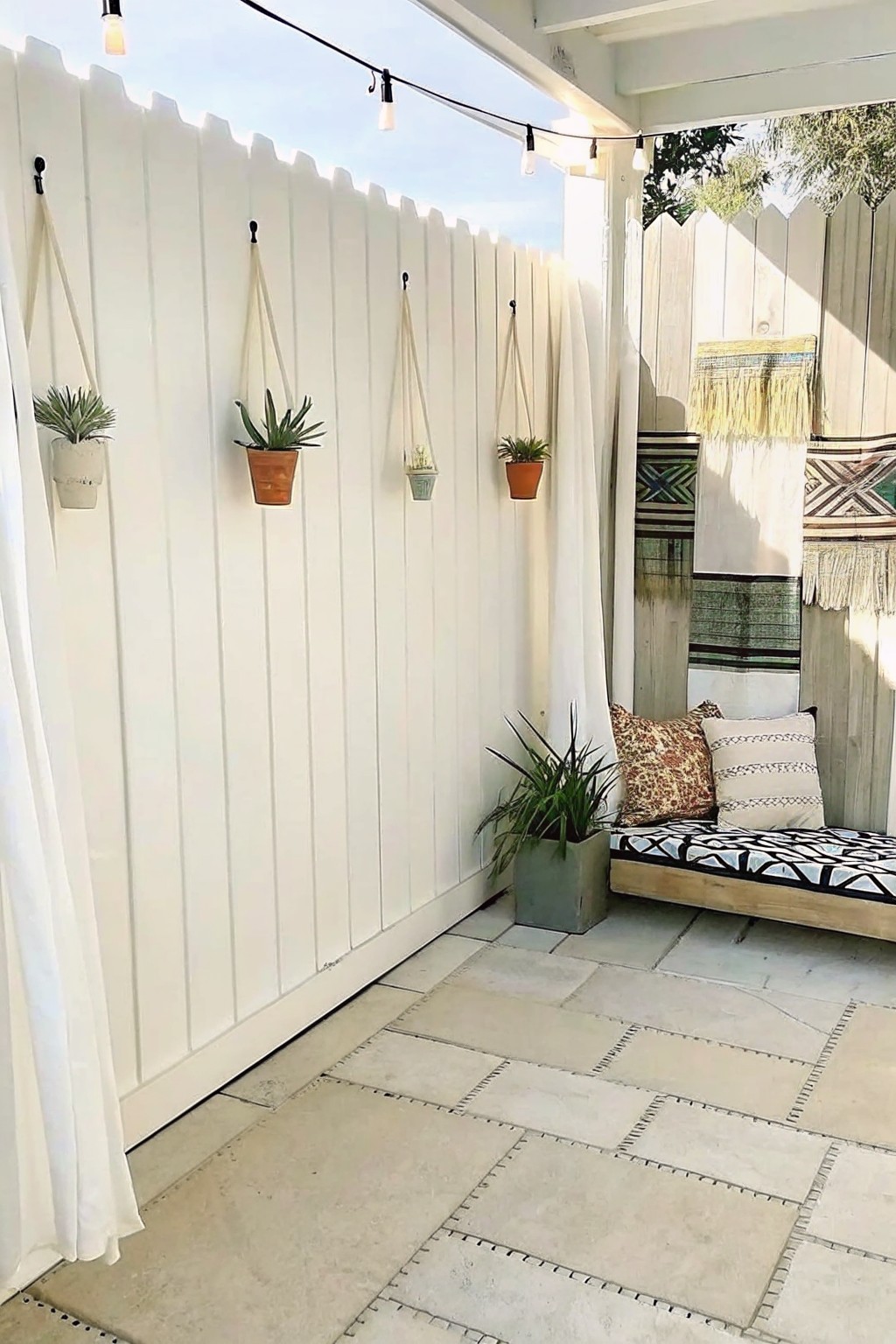 A white fence with hanging potted plants above a paved patio area that includes a low daybed with pillows.
