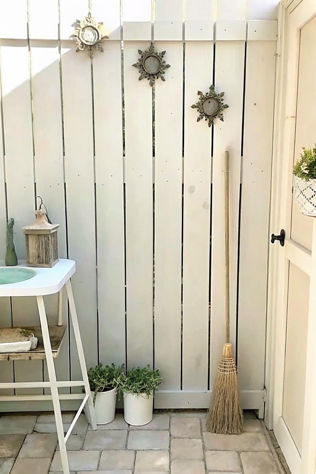 Whitewashed plank wall with sun ornaments, broom, potted plants, lantern, and stone floor