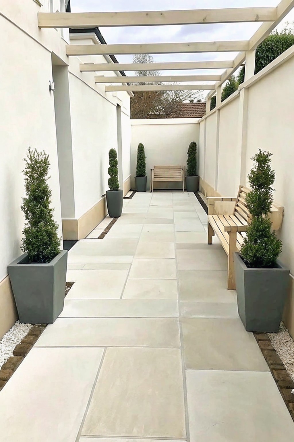A narrow paved outdoor walkway between tall white walls, lined on both sides with gray rectangular pots holding tall conical evergreen shrubs, wooden benches, and a light wood pergola overhead.