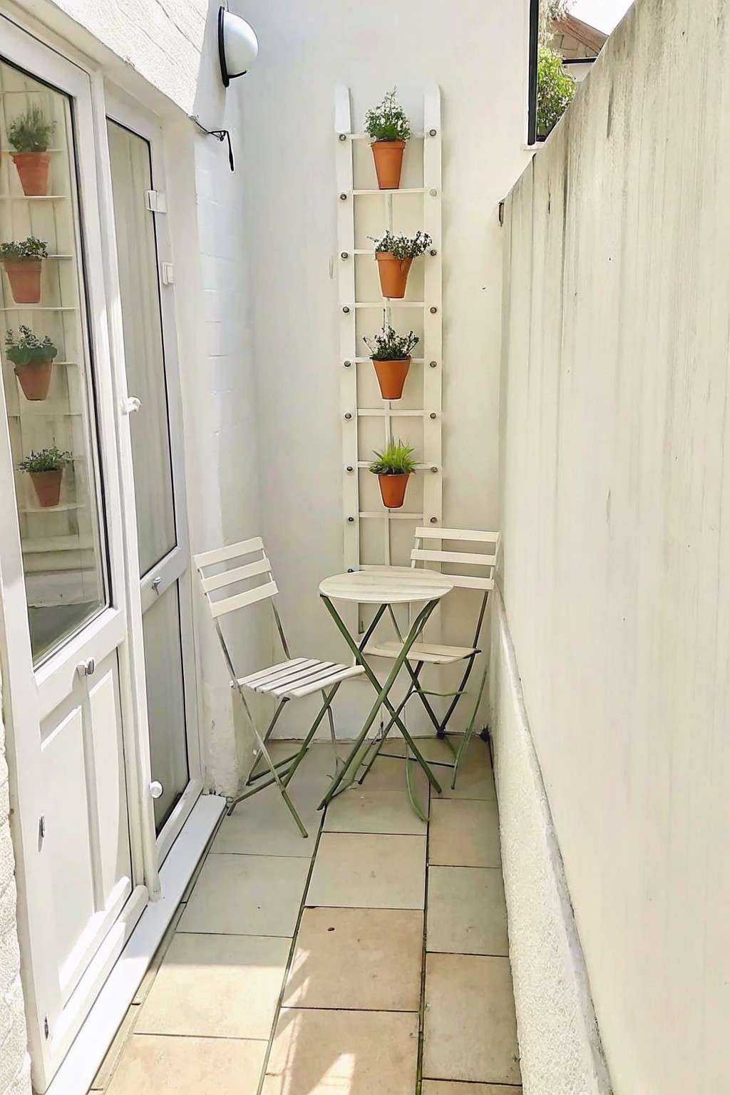Narrow outdoor walkway with a white vertical ladder holding potted plants on the wall beside a small round table and two white folding chairs.