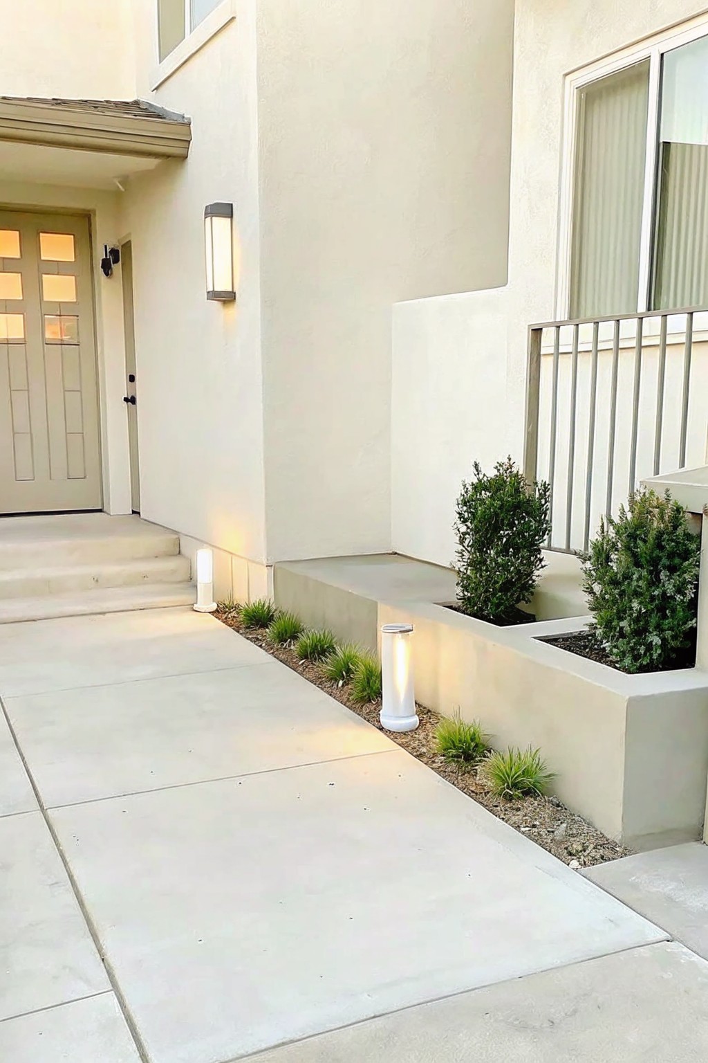 Concrete walkway next to a house with a gravel border of low plants, two cylindrical lights, and raised concrete planters holding shrubs.