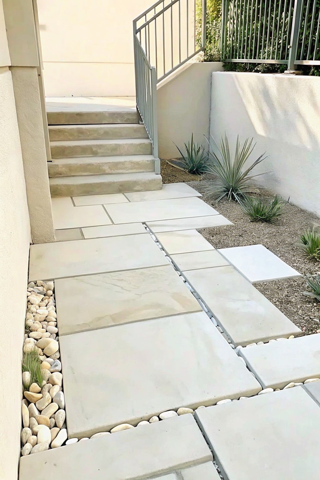 Modern stone paver walkway with white pebble borders beside concrete stairs and metal railing