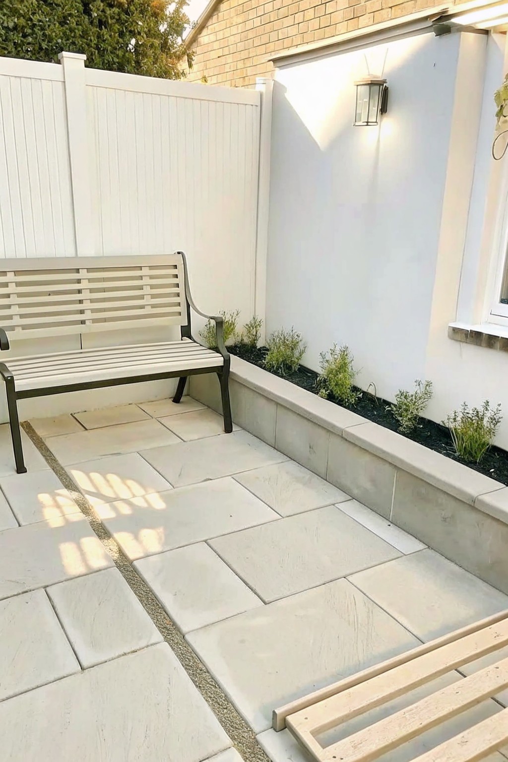 A stone patio beside a long raised concrete border with small shrubs planted in a narrow bed along a white wall, and a white bench on the left.