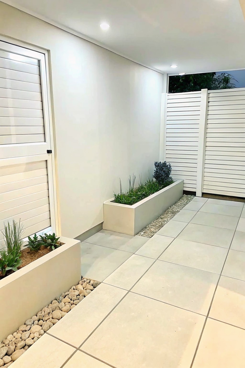 A light-tiled outdoor walkway bordered by rectangular concrete planters filled with green plants and edged with light gravel, next to a white slatted fence.