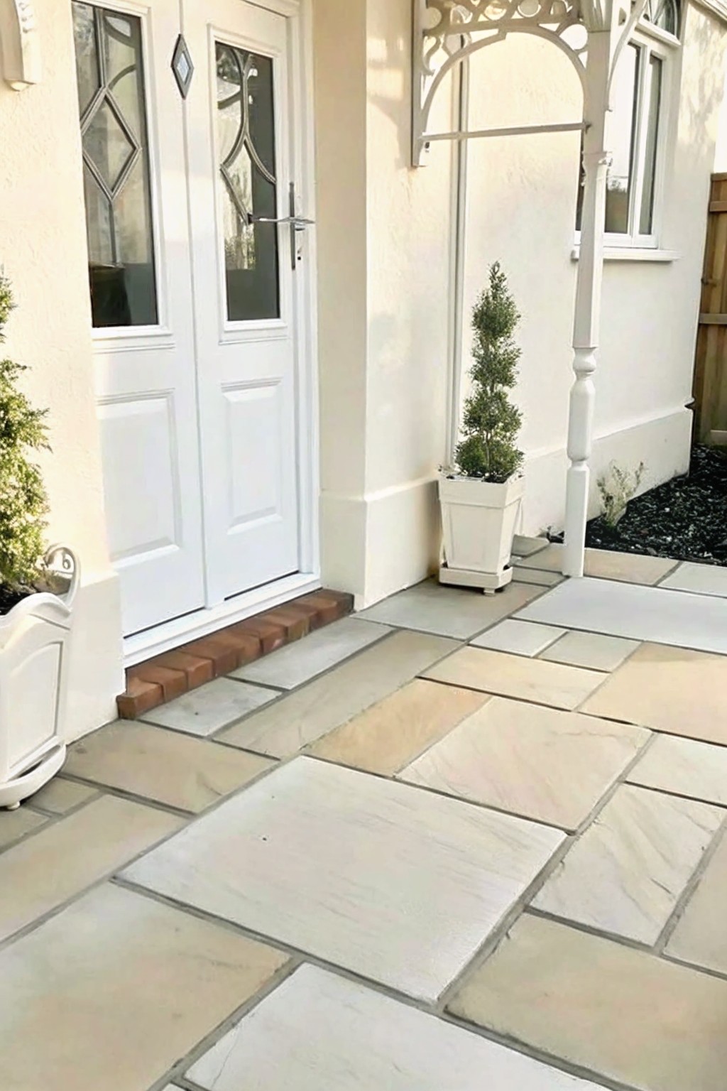 A patio of large light-colored stone pavers leads to white double doors, with a potted evergreen shrub placed along the wall.