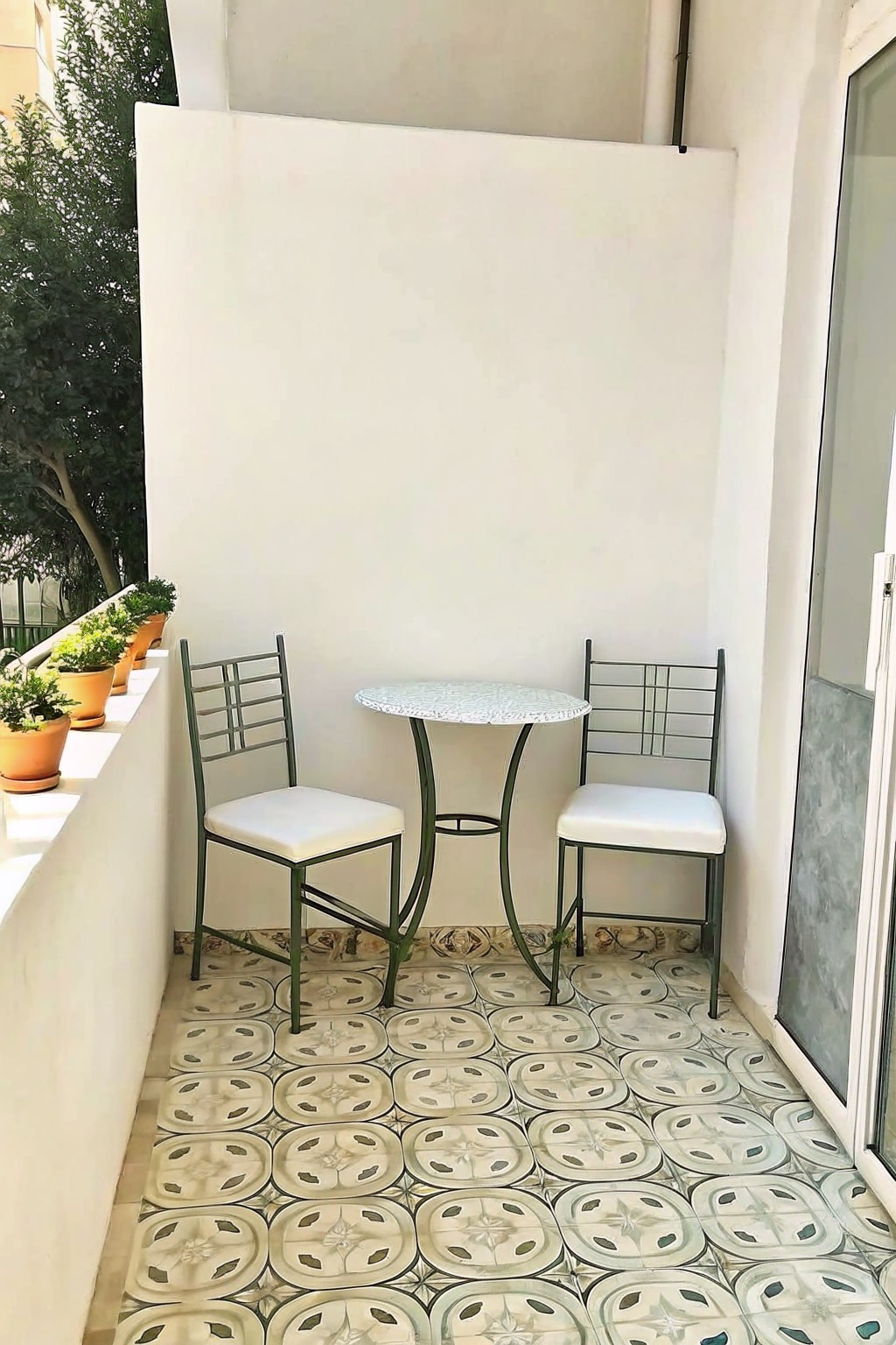 A small balcony corner with a round table and two chairs on patterned tiles, white walls on three sides, and potted plants along the left ledge.
