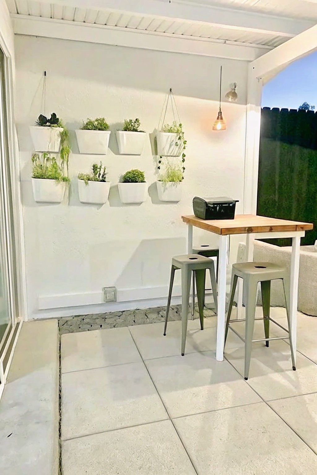 An outdoor patio with a white wall holding multiple rows of white rectangular planters filled with green plants, a wooden table with two metal stools, and a hanging pendant light.