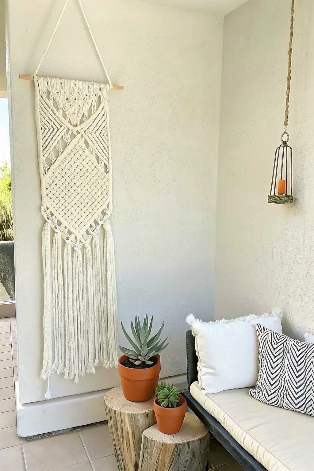 A large off-white macrame wall hanging on a textured exterior wall beside a cushioned bench and two potted succulents on wooden stumps.