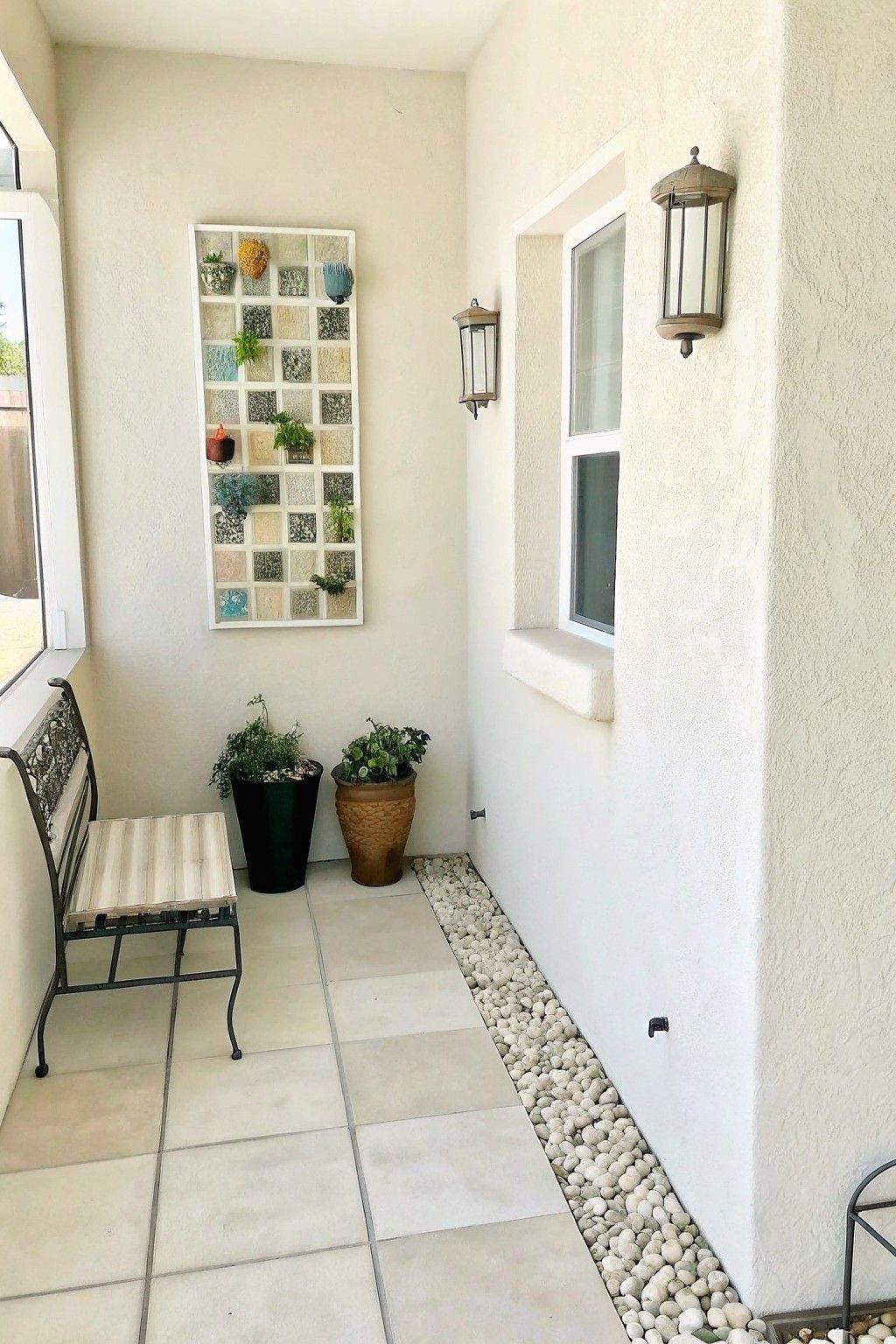 A light-colored patio wall features a white rectangular grid frame filled with small potted plants above two larger pots and a metal bench.