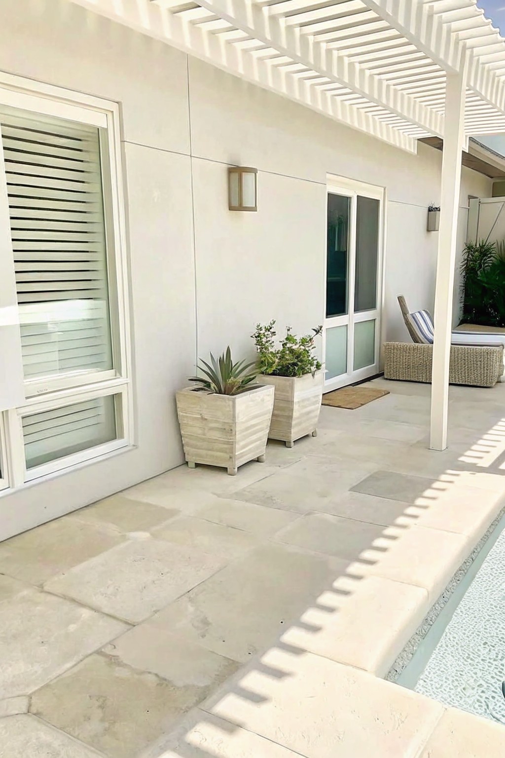 White patio with stone tiles, two planters, pergola, and poolside lounge chairs.