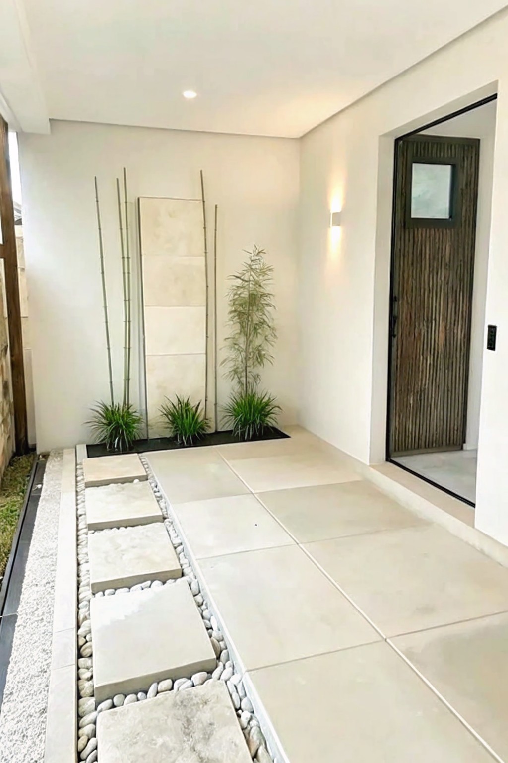 Minimalist entryway with large stone tiles, pebble path, bamboo plants, and wooden door.