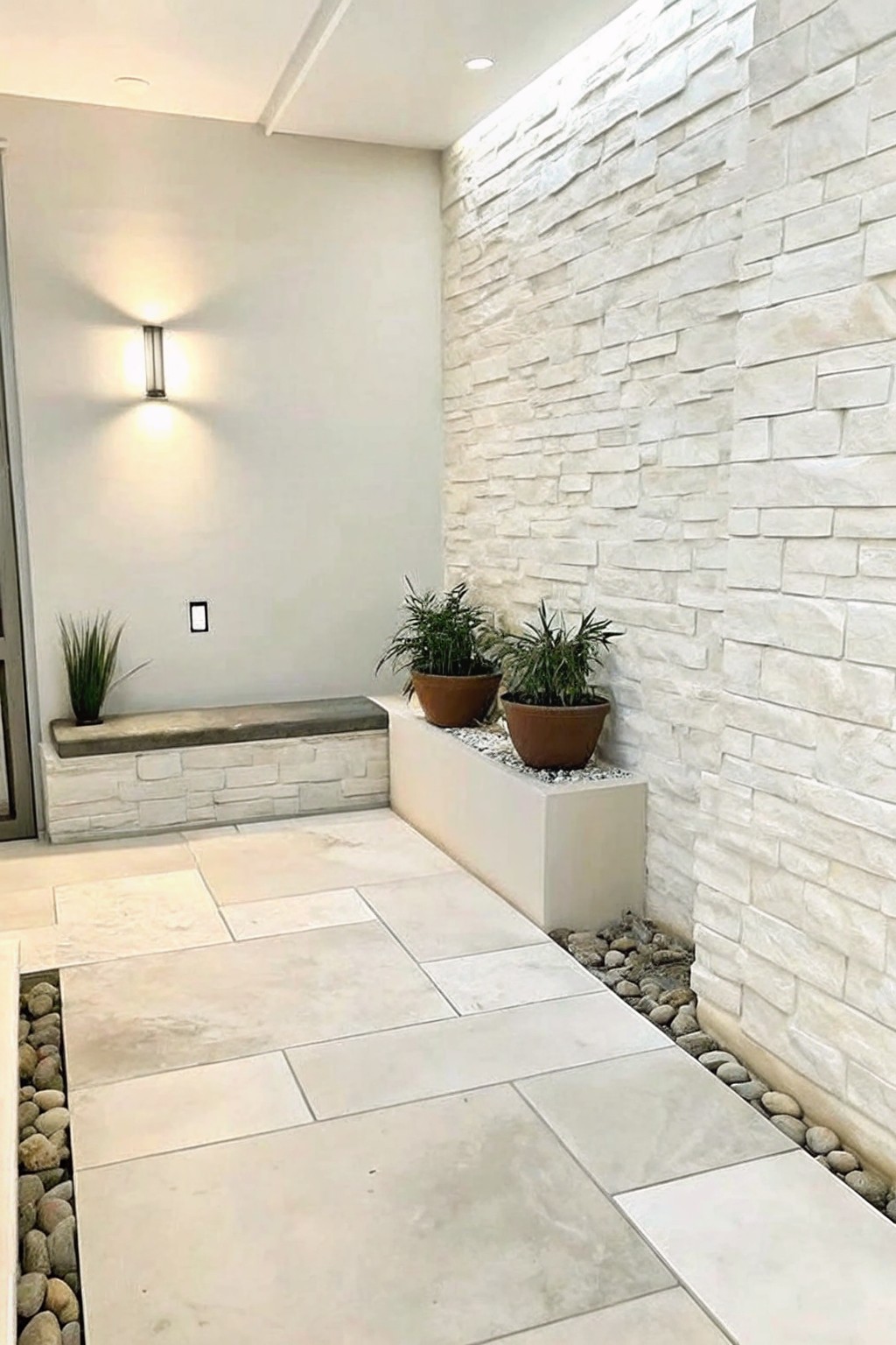 A compact outdoor space with large light tiles, pebble borders, a white stacked stone wall holding two potted plants on a low ledge, and a built-in bench against a plain wall.