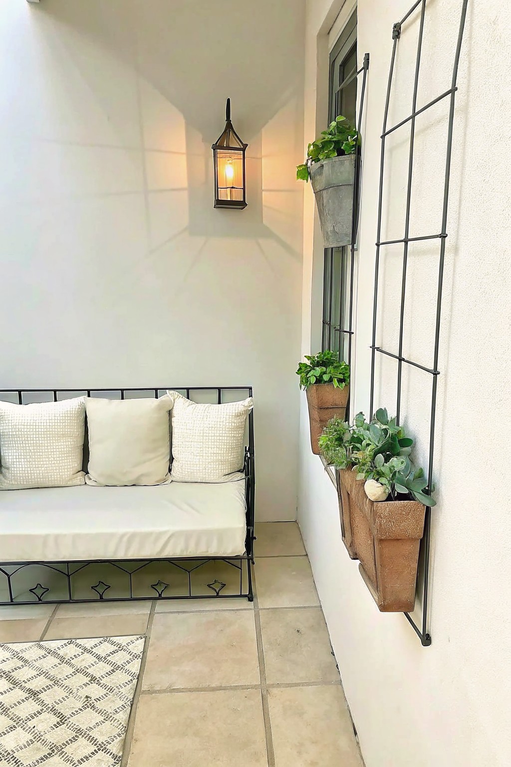 An outdoor bench with cushions sits against a white wall beneath a lit black lantern, with vertical metal planters mounted beside a window on the right.