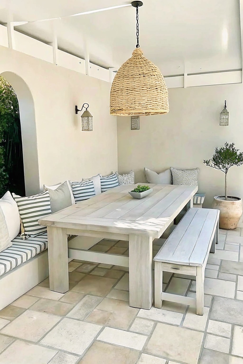 A covered outdoor patio featuring a long wooden dining table, built-in cushioned seating along three sides, a matching bench on the fourth side, and a large woven pendant light suspended above.