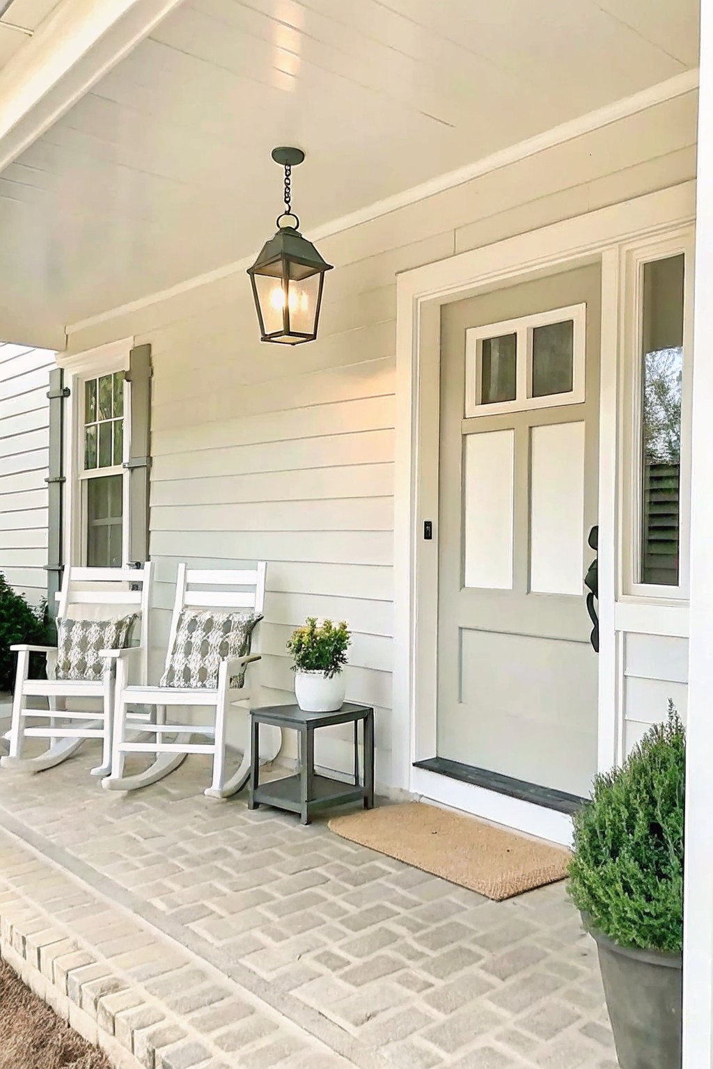 A front porch with two white rocking chairs, a small gray side table holding a potted plant, a hanging lantern, and a light gray door beside a doormat.