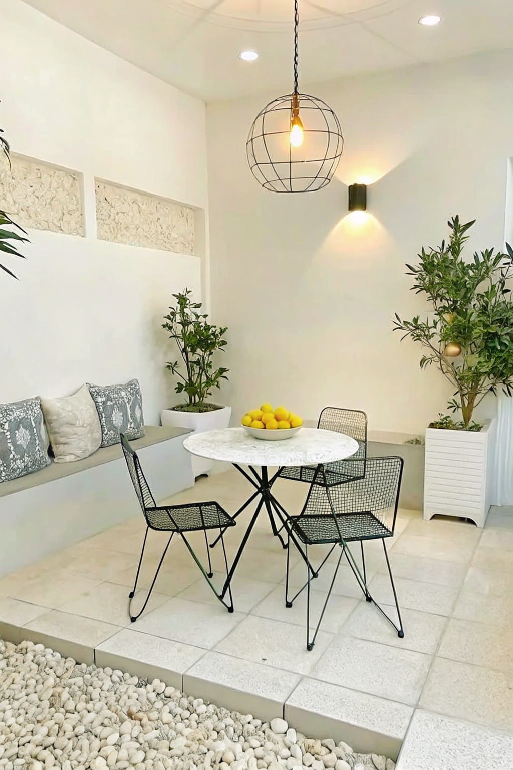 An outdoor patio with a round marble table, black wire chairs, a hanging spherical pendant light, potted plants, and a built-in bench with cushions.