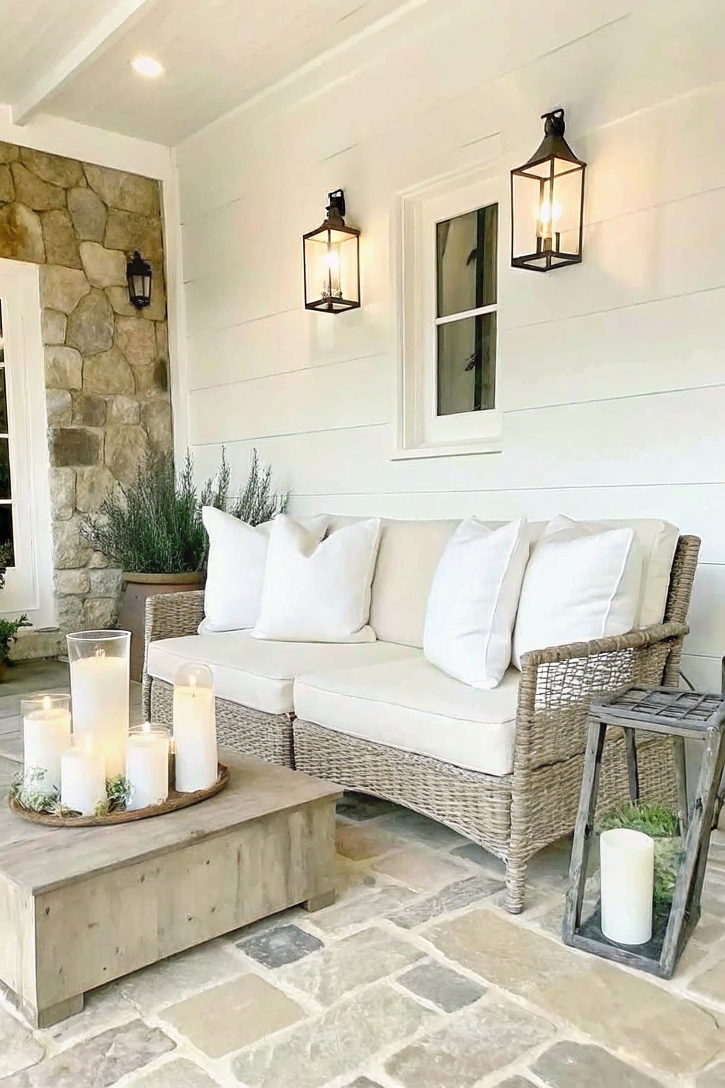 Patio with wicker sofa, white cushions, wooden table of candles, and wall lanterns.