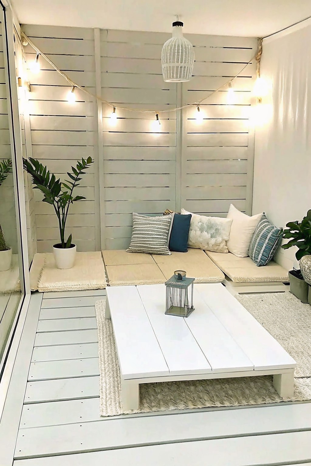 Outdoor deck with low white platform seating, beige floor cushions and pillows, string lights along white walls, potted plants on the sides, and a white coffee table holding a small lantern.