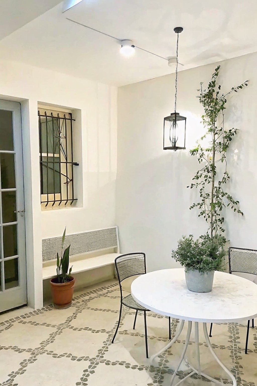 White courtyard with round marble table, mesh chairs, plants, lantern, and patterned floor.