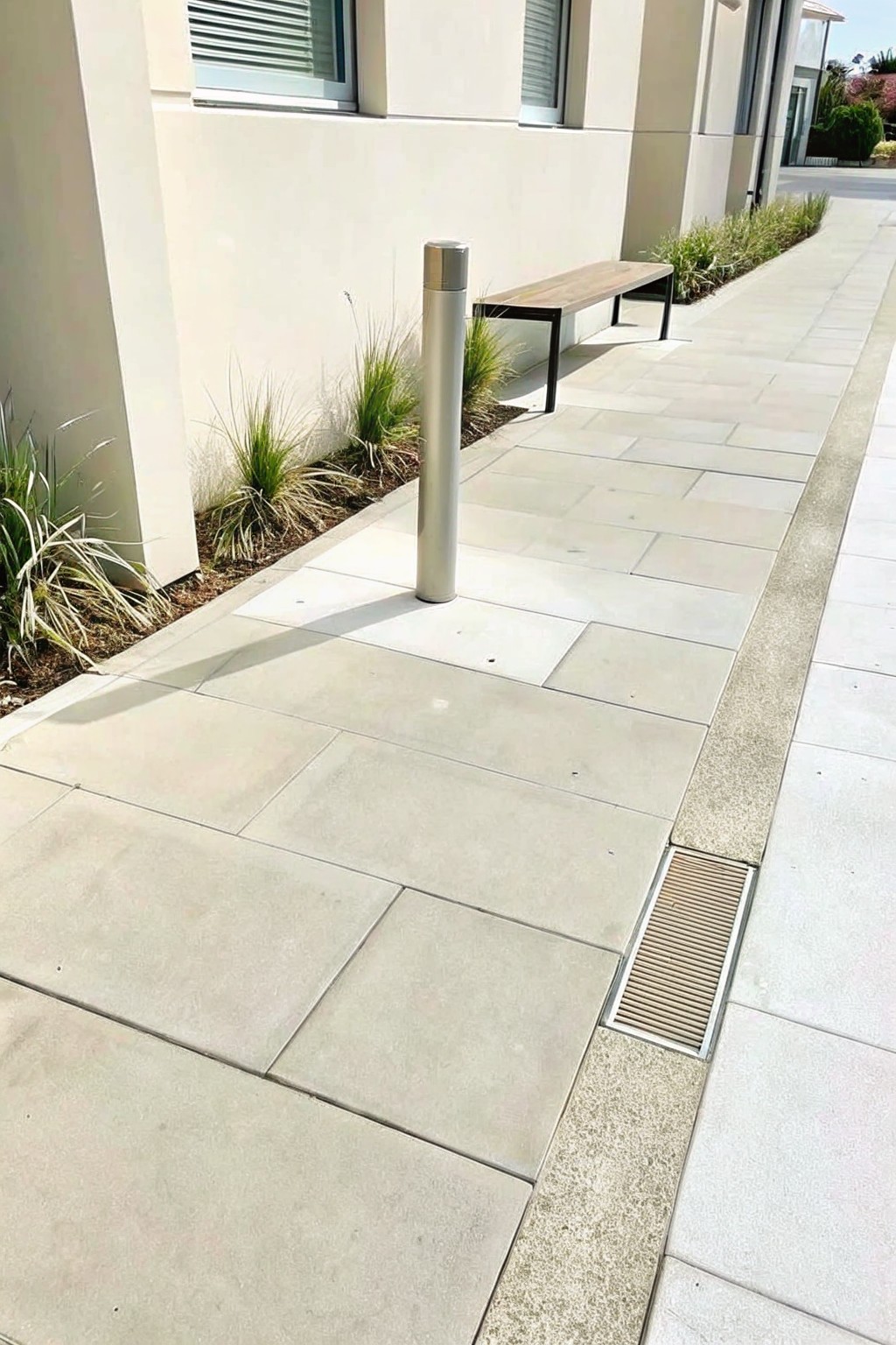 Stone-paved walkway beside beige building with metal bollard, wooden bench, and drainage grate.