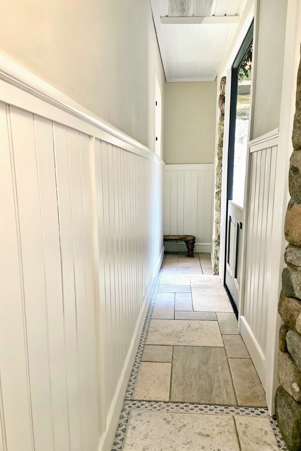 A narrow interior hallway with white wainscoting on both walls, a tiled floor with a decorative pebble border along the edges, and an open door showing a stone wall outside.