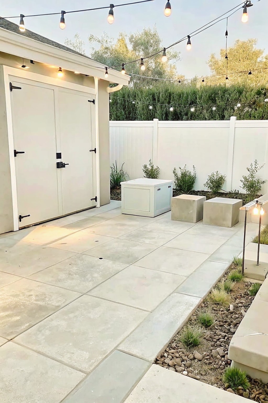Modern backyard patio with white doors, string lights, concrete seating, and garden beds