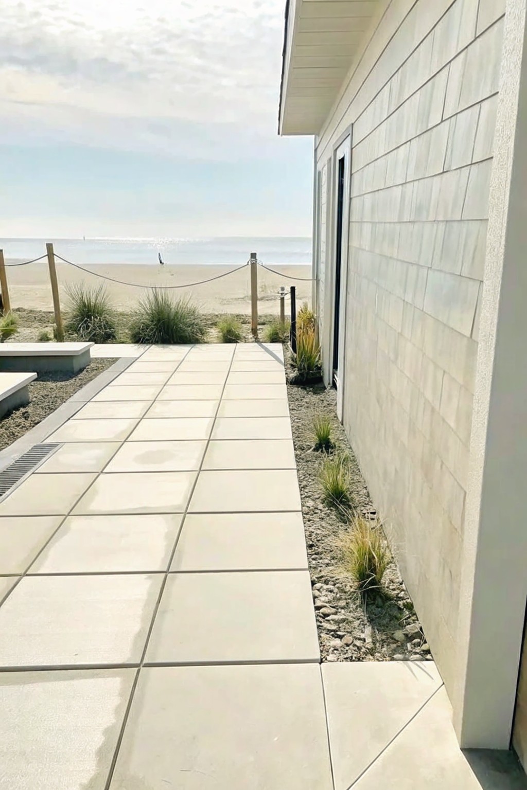 A light-colored paved path runs next to a house, with a narrow gravel strip and clumps of grasses bordering the edge between the path and the wall.