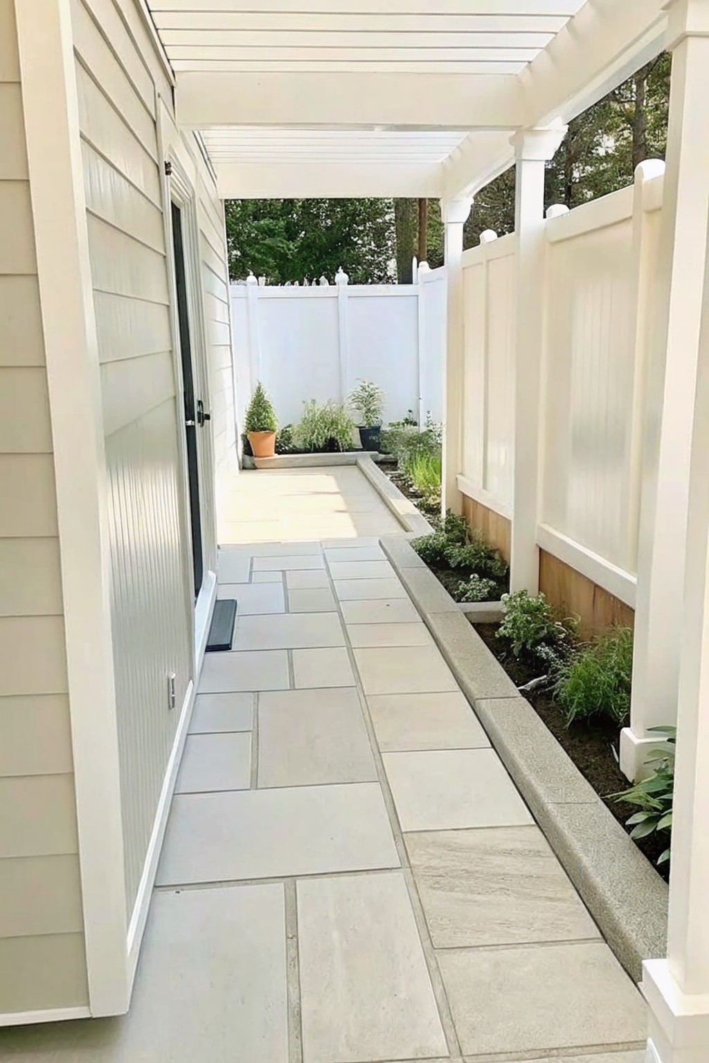 Covered stone walkway under white pergola beside house and garden bed
