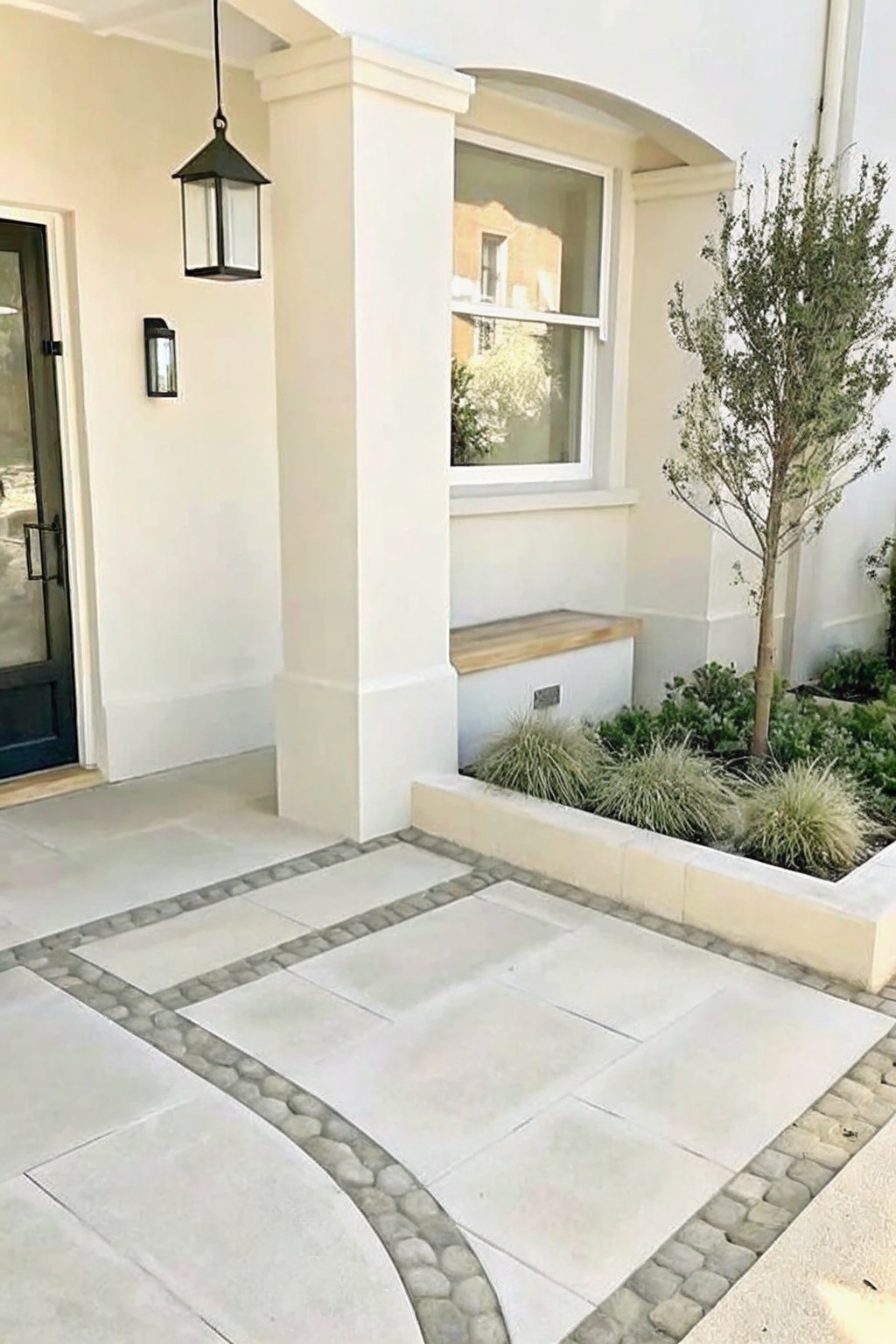 A stone-paved walkway bordered by cobblestones leads to a raised planter bed with grasses and a small tree beside a house entrance.