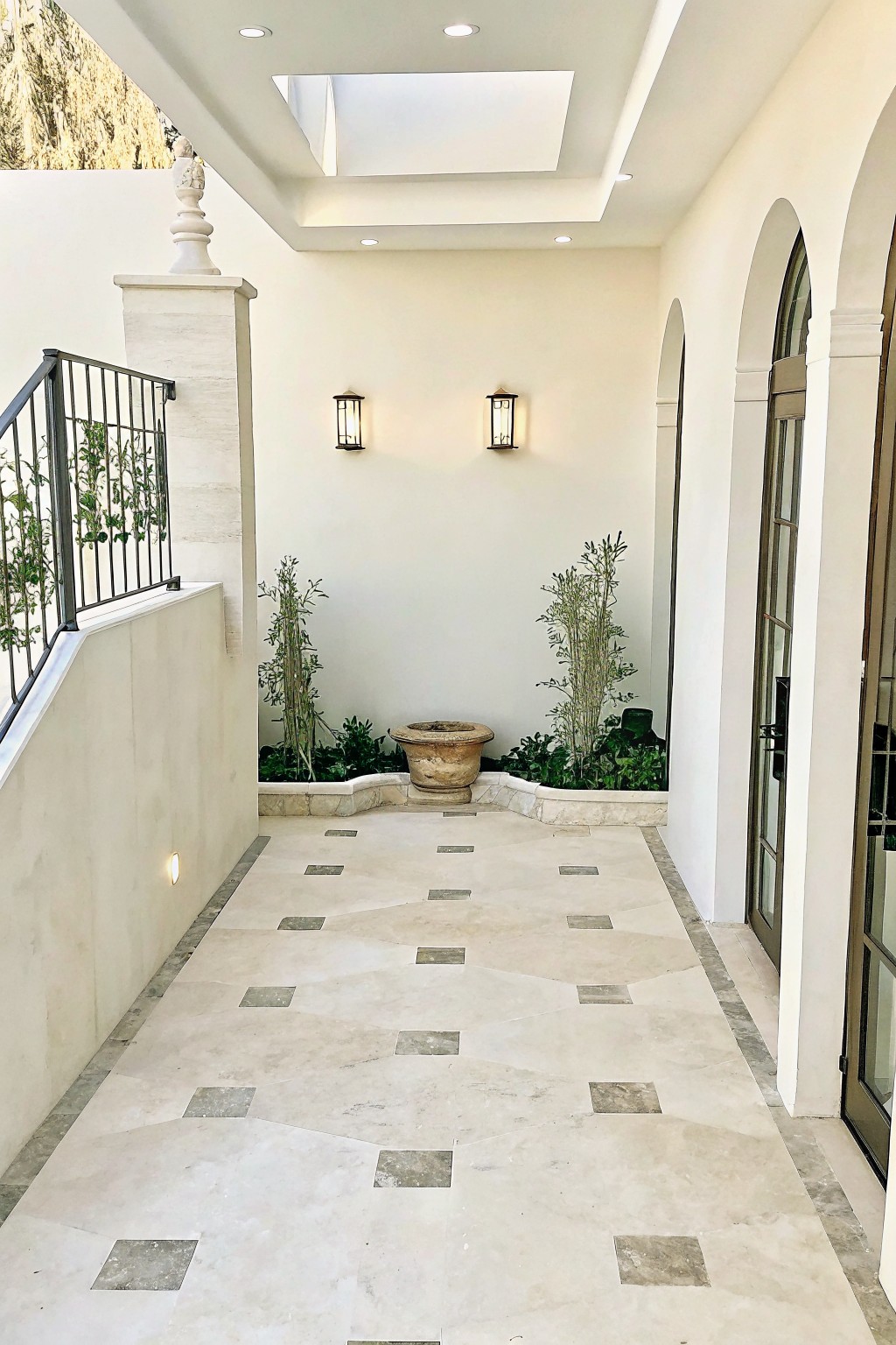 A covered outdoor walkway with light tile flooring shows raised planting beds edged in light natural stone and filled with dense greenery and a stone urn.